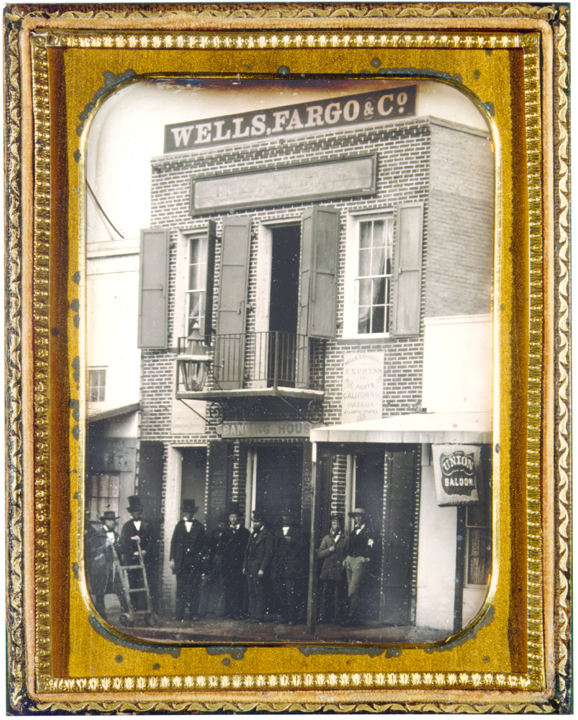 Photo in gold frame shows a two story building brick building with sign Wells Fargo & Co. A half-dozen men in business attire stand outside open doorway. Image is black and white in gold gilded frame.