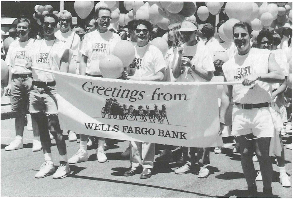 Group of men and women in white t-shirts marching in parade behind horizontal banner reading Greetings from Wells Fargo Bank. Banner also has logo of stagecoach and running horses. Festive balloons in background.