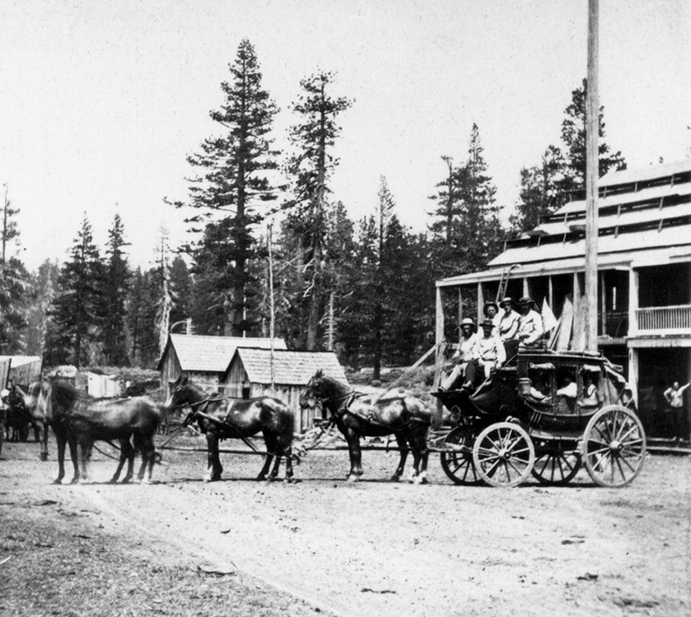 Stagecoach drawn by six horses faces left on dirt road outside wooden building. Five men are on top of coach, including driver, and several passengers inside. Forest of tall pine trees in background. Image is black and white.