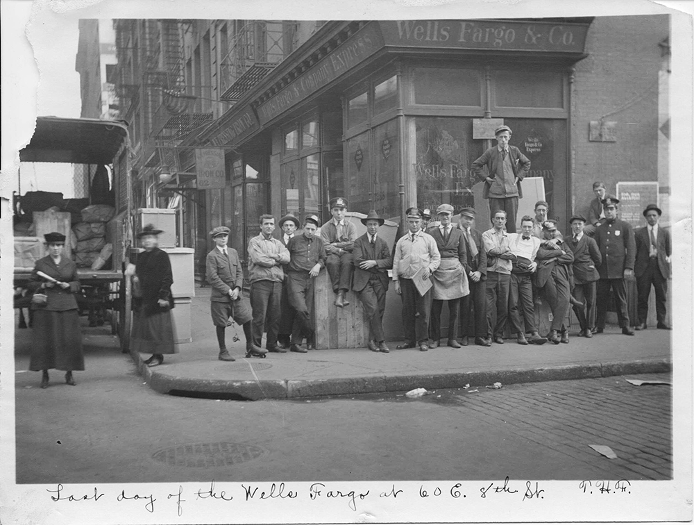 Group of working men and women casually posed standing gathered on sidewalk outside of building in New York City. Building has sign reading Wells Fargo & Co's Express. Loaded wagon at left. Handwritten caption reads Last Day of the Wells Fargo at 60 E 8th Street. Image is black and white.