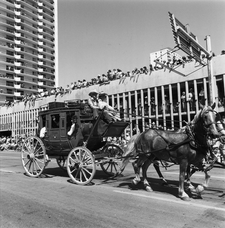 Multiple people ride on top of a stagecoach pulled by horses down a city street. Only two horses are shown. Several passengers wave from inside of stagecoach. Parade viewers crowd low rooftops in background. Tall city building at left background. Image is black and white.