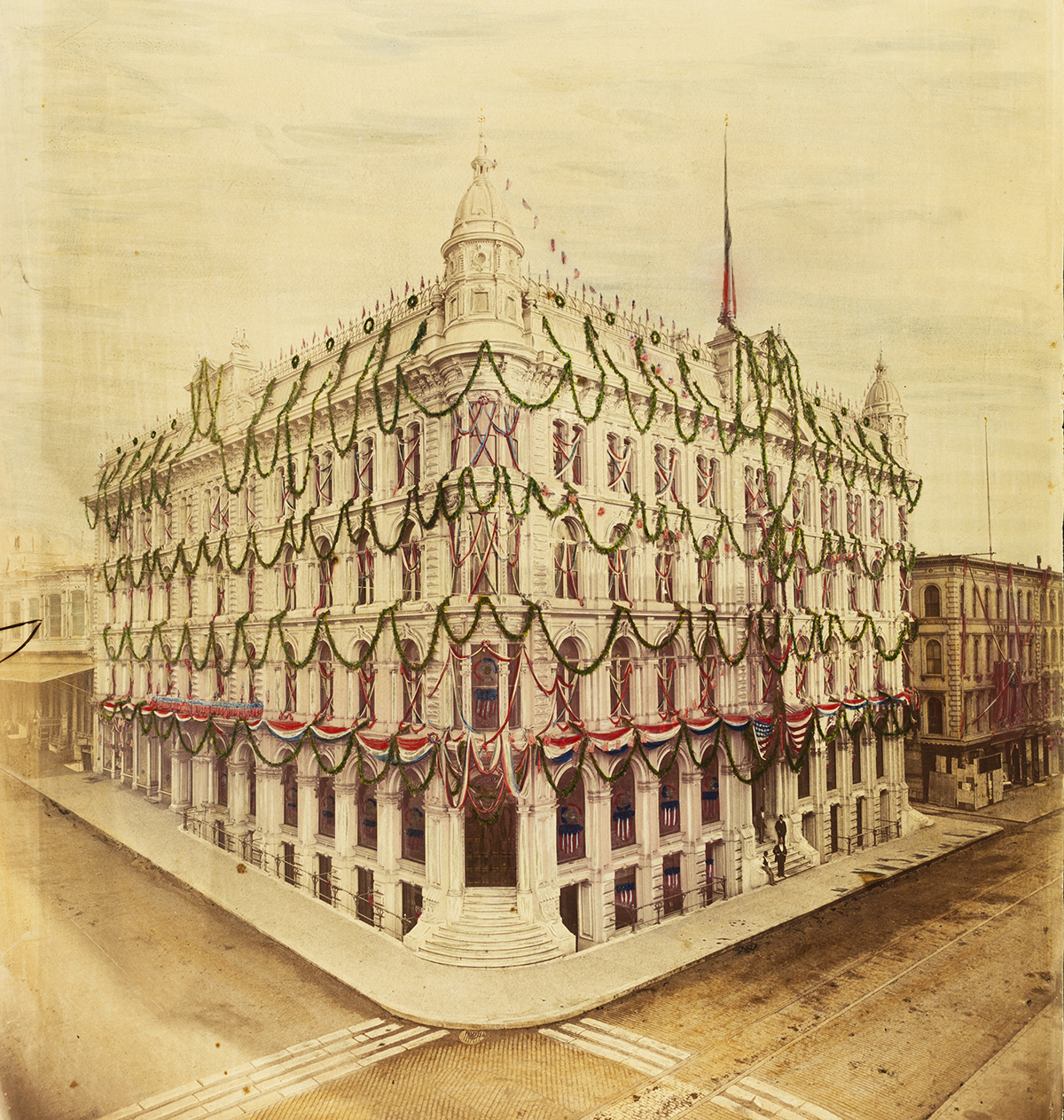 Hand-colored photo of a four-story bank building on corner. The building has several domes and finials on its ornate rooftop, and is decorated with patriotic bunting, tinted in red white and blue. Photo is a yellowed black and white image.
