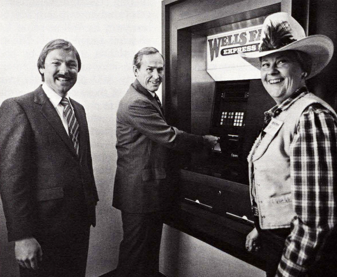 Two Caucasian men in suits stand in front of ATM machine labeled with name Wells Fargo. One man looks at camera while second man uses ATM. A Caucasian woman stands to right of machine turned toward camera. She is dressed in western style vest, shirt and hat. Image is black and white.