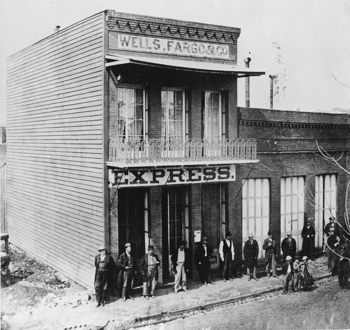 Historic photo of two-story brick building with large sign at top reading Wells Fargo & Co., and above the door a second large sign reading Express. A crowd of men, women and children are gathered outside. Image is black and white.