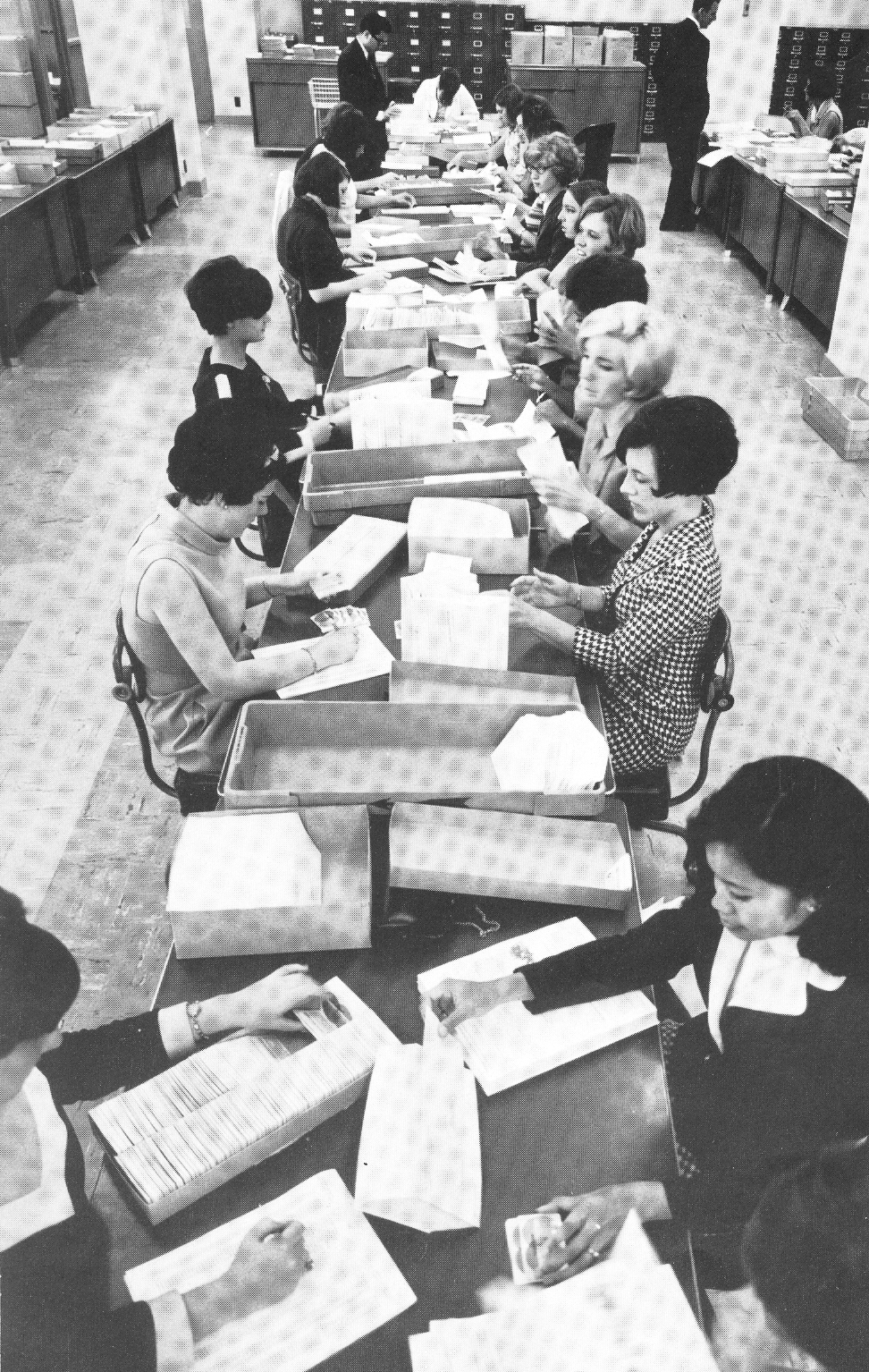 A group of women sit at long table stuffing envelopes with credit cards and letters. Boxes of mailing materials sit on the table. Image is black and white.