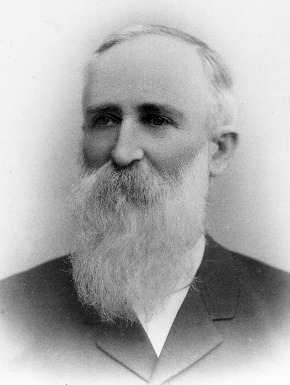 Head and shoulders portrait of Caucasian man with long white beard. Man is dressed in dark suit, looking slightly right. Image is black and white.