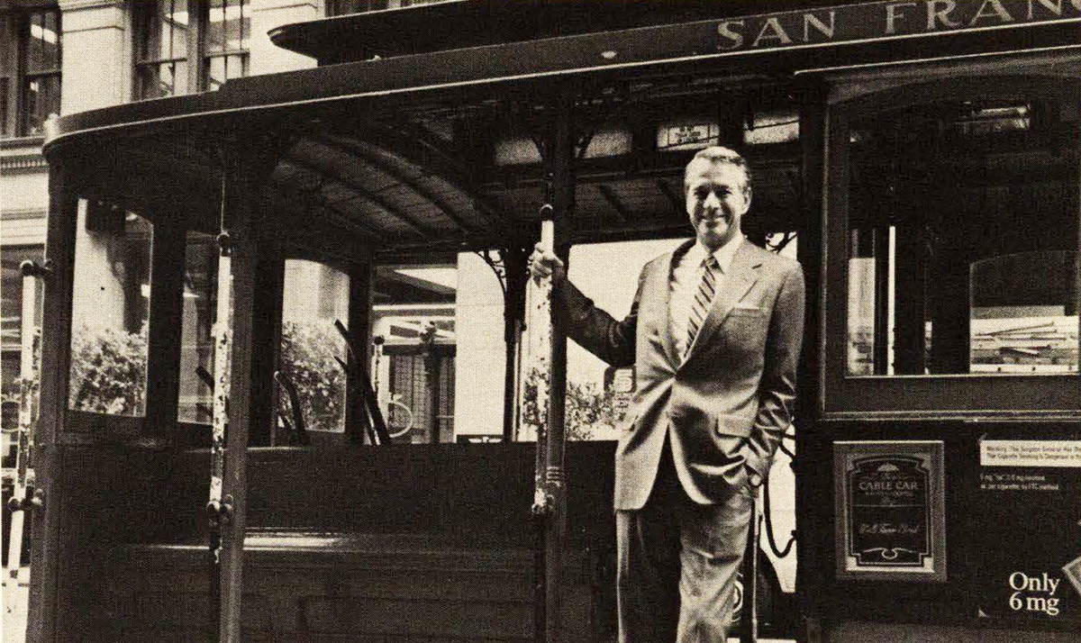 Caucasian man in a suit stands aboard San Francisco cable car, facing camera. Black and white image.
