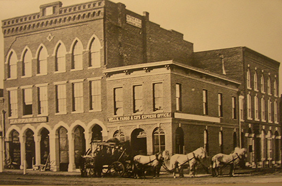 Stagecoach and six harnessed horses stand outside two-story brick building with sign reading Wells Fargo & Co.'s Express office. Building with sign is on street corner, other commercial buildings either side. Image is black and white.