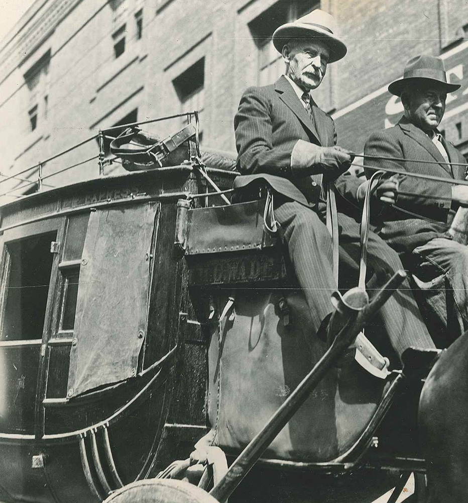 An elderly Caucasian man in suit and tie holds reins in both hands while driving a stagecoach. A second man sits to his left. No horses are shown. Photo is taken from street level, looking slightly up. City building in background. Photo is in black and white
