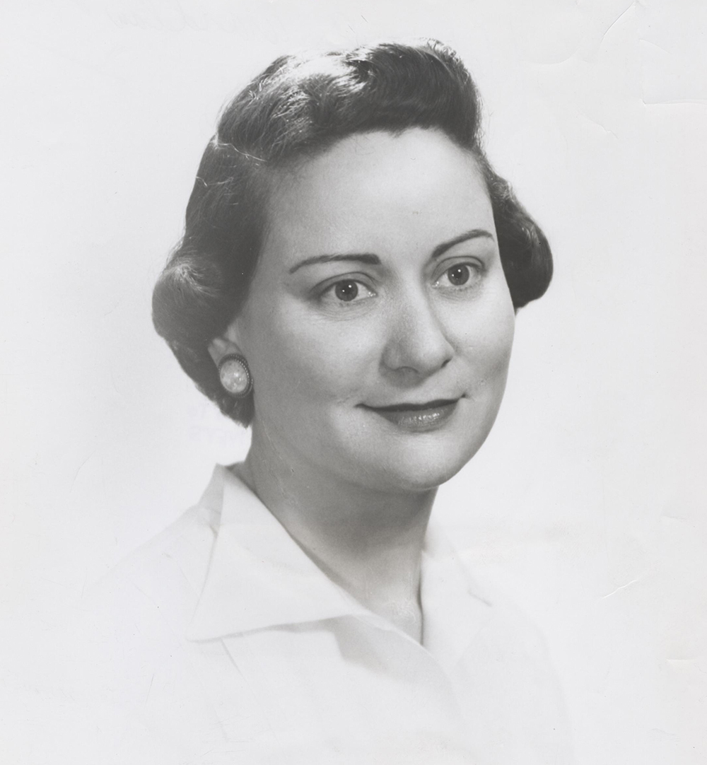Head and shoulders portrait of Caucasian woman looking left of camera. She is dressed in business attire white blouse. Her dark hair is neatly combed back, she wears large white earrings and dark lipstick. Image is in black and white.