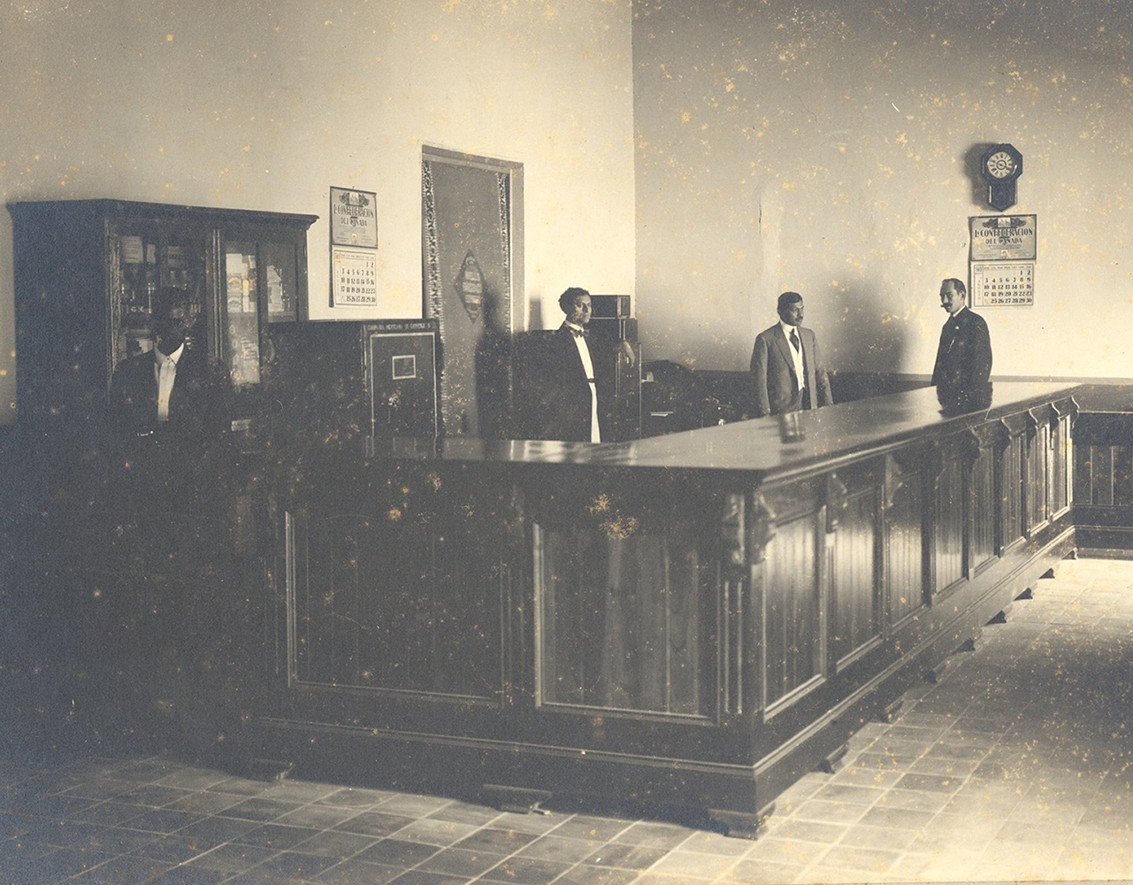 Photo of office interior where four men stand in room behind dark wood counter, facing camera. All are dressed in suits. Bookcase, clock, and wall calendars in background. Image in black and white.