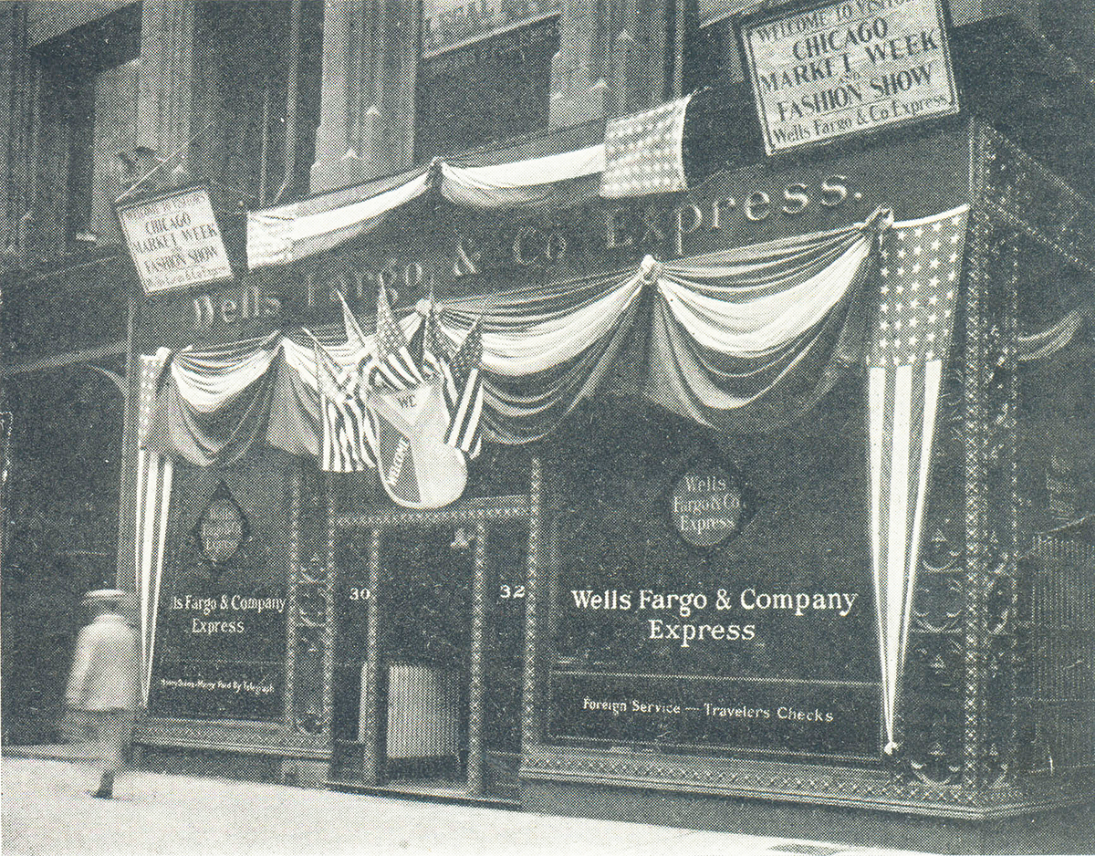 Building front draped in patriotic bunting above large glass windows. Sign reads welcome visitors Chicago Market Week and Fashion Show and Wells Fargo & Company Express. Person walks right to left facing away on sidewalk in front. Image in black and white.