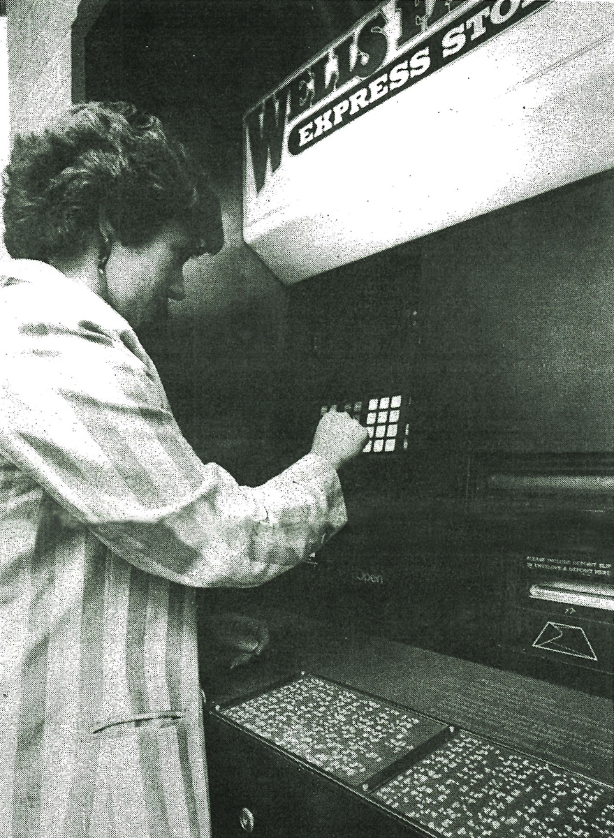 Woman wearing striped jacket stands to left in front of ATM machine. Her right hand pushes buttons on machine. Raised braille panels are visible beneath machine display screen and keypad. ATM signage reads, in part, Wells Fargo Express Stop. Image in black and white.