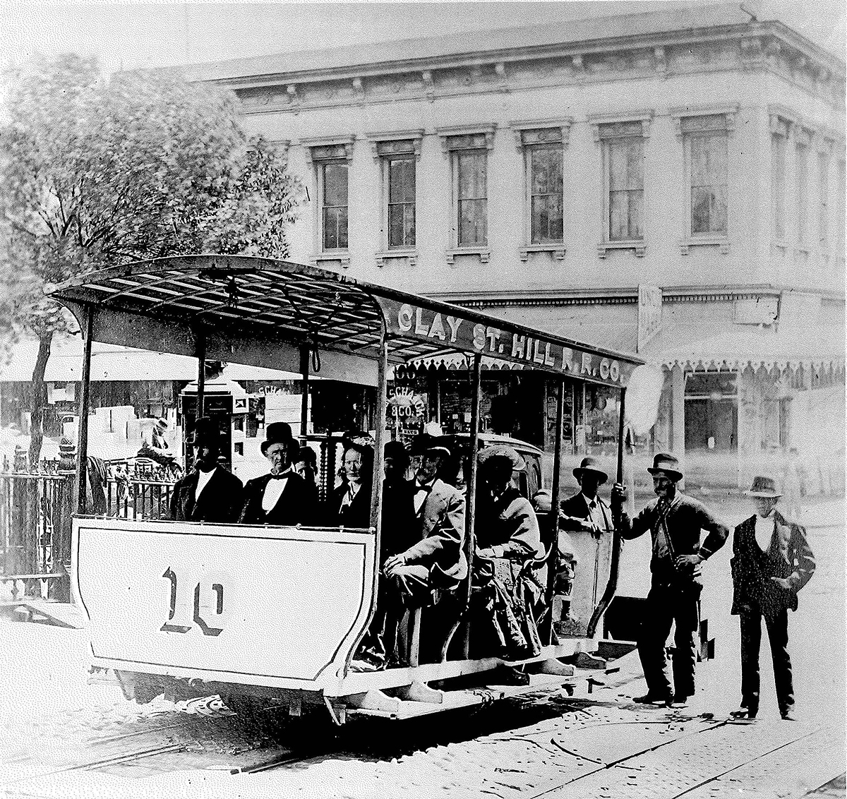 A short vehicle marked number ten is stopped on tracks climbing a hilly street. Well-dressed men and woman passengers sit facing forward on three bench seats in open-air cable car labeled Clay Street Hill Railroad Company. Two men stand behind the car. Building and trees in background. Image is black and white.