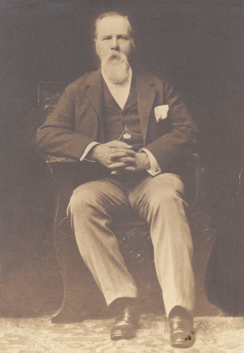 Studio photo portrait of seated Caucasian man with gray hair and long beard. His hands are clasped in his lap. He is wearing a dark suit and tie, lighter pants and black shoes. Image is black and white.