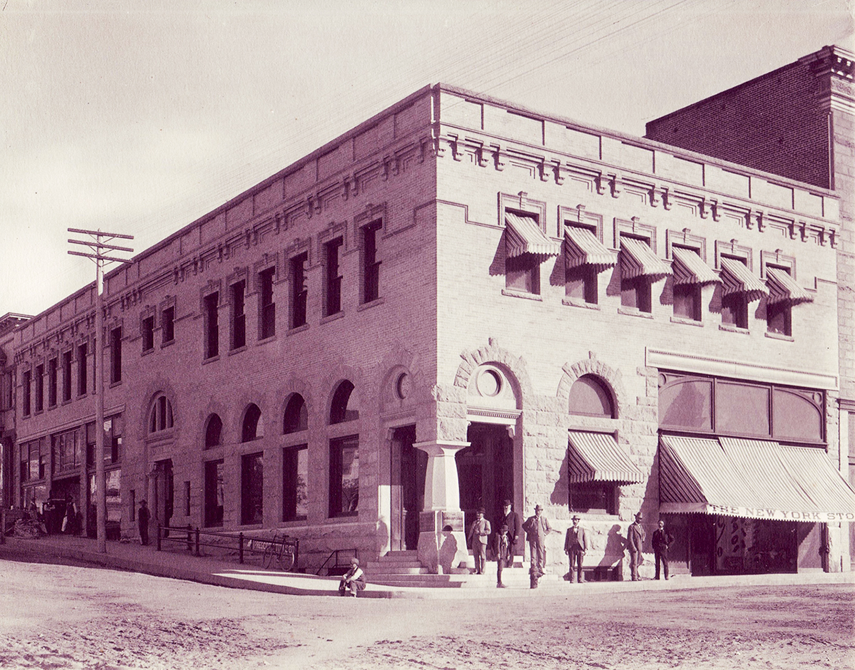 Two-story stone building on street corner, several men pose outside the door, which is located at the corner of the building. Image is black and white.