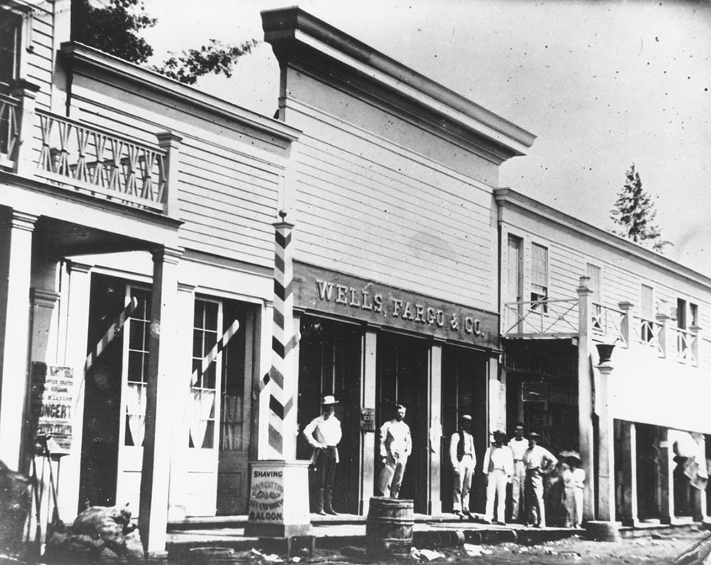 Streetview of three wooden single-story storefront buildings. Center building has sign reading Wells, Fargo & Co. overhead. Several men stand outside on wood plank sidewalk. Barbershop sign shown next door. Image is black and white.