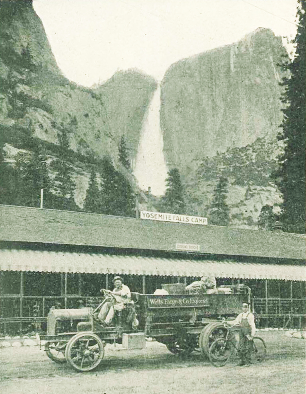 Man drives truck full of luggage and packages. Wells Fargo & Co Express sign in bed of truck. Second man in work clothes stands with bicycle at rear of truck. Building in background with Yosemite Falls waterfall filling mountain background in distance. Image is black and white.