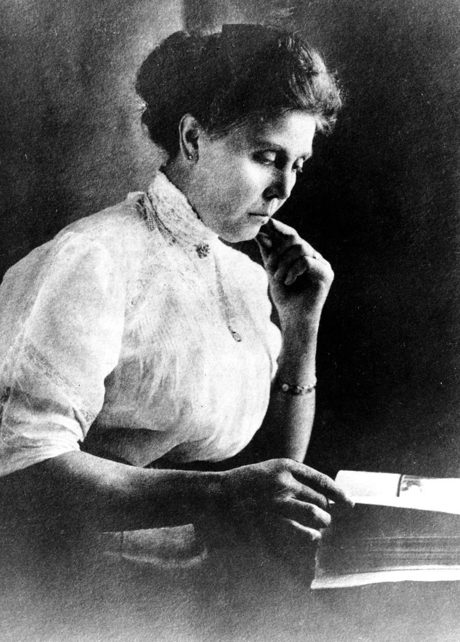 Woman sits turning pages of book open on tabletop. Her right hand turns page while her left elbow rests on table and left hand is raised to her chin. Her dark hair is pinned on top of her head, and she wears a bright white blouse. Image black and white.
