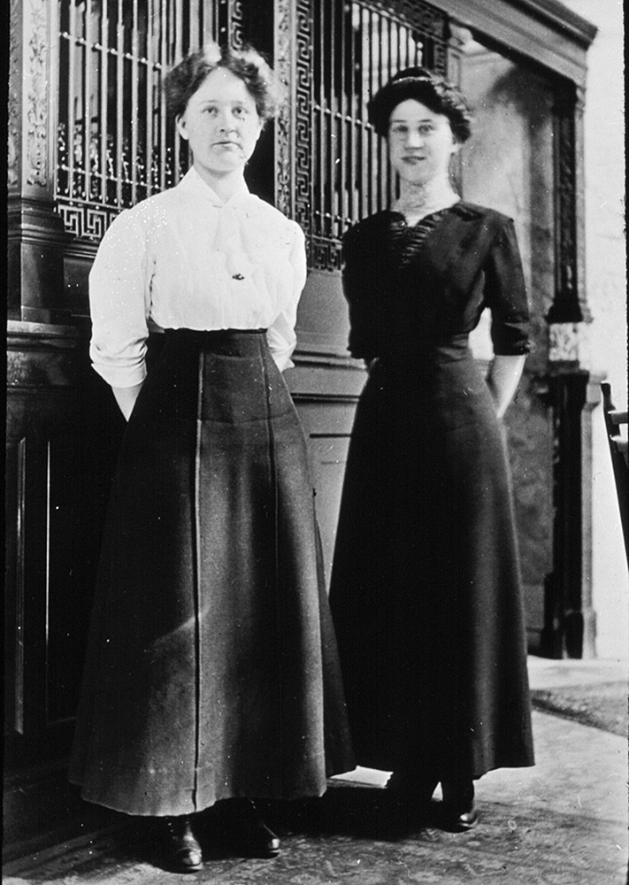 Two women pose side-by-side standing in front of bank teller window, arms clasped behind them, hair neatly pinned up. Woman at left wears long skirt and white blouse, woman at right dark floor-length dress. Image is black and white.