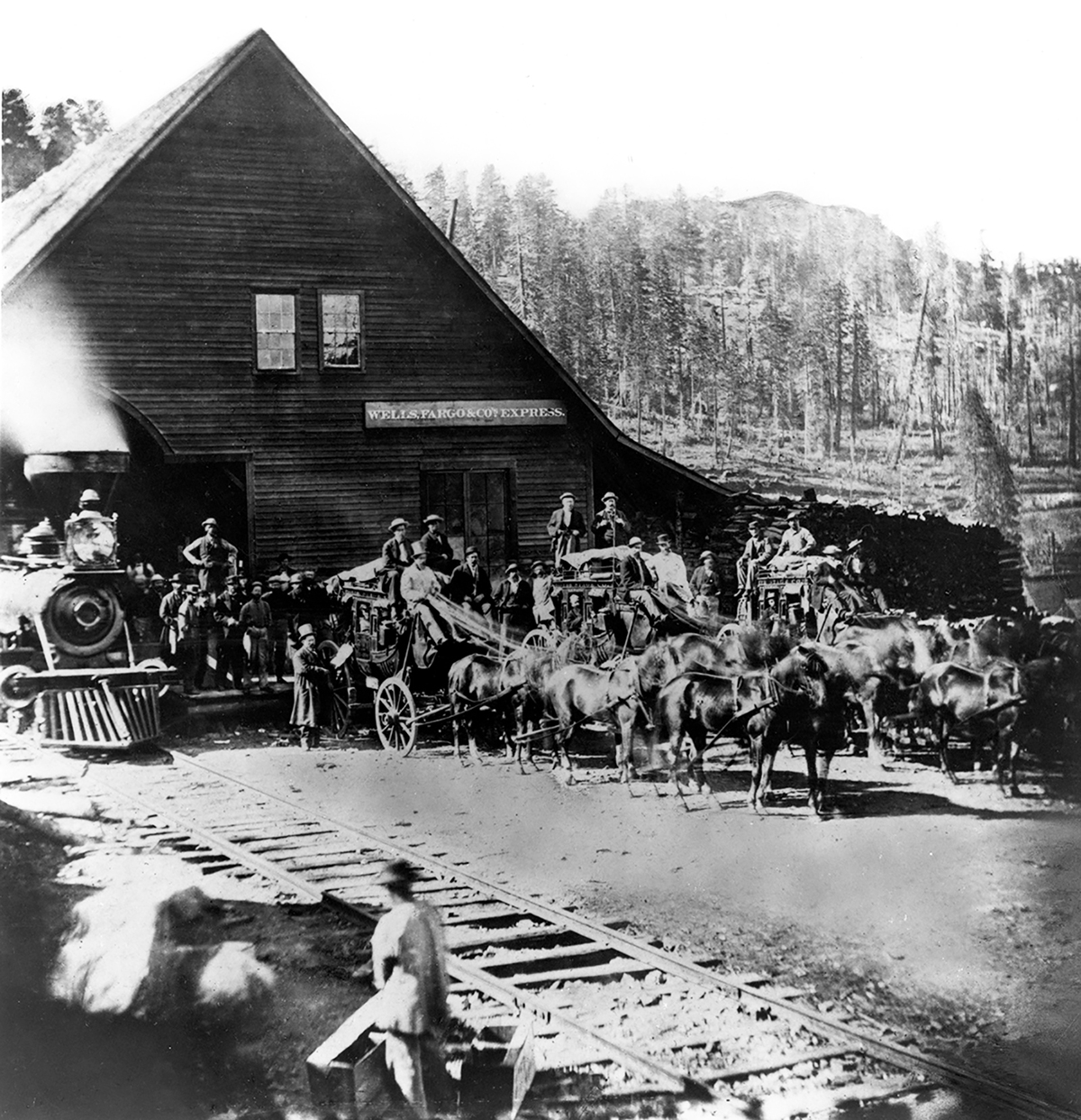Peak-roofed wooden building, forest in background. Steam train approaches from left. Three stagecoaches stand next to building under sign reading Wells Fargo & Co' Express. Stages are loaded with passengers and hitched to horses. Image in black and white.