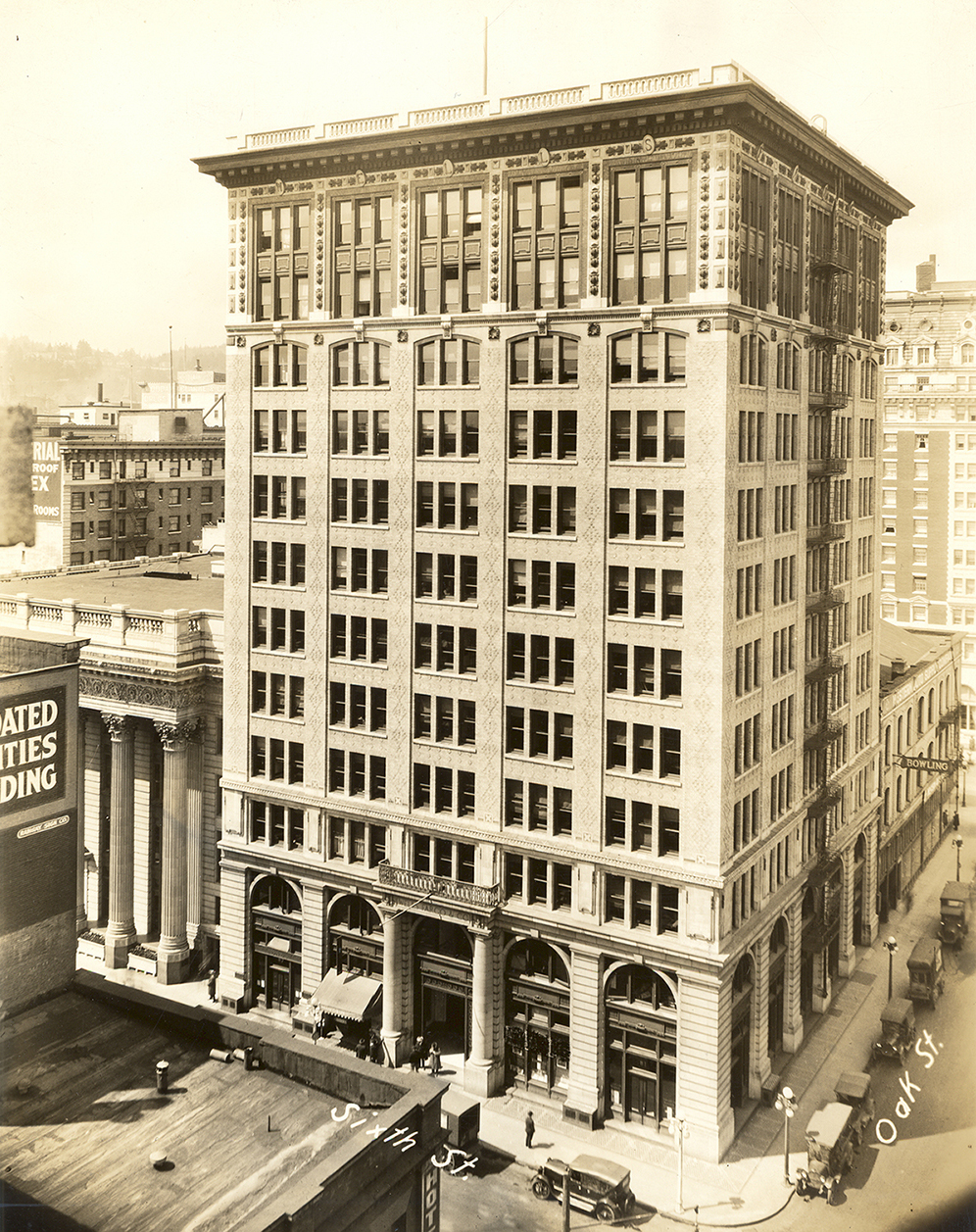 Photo of tall building on street corner. Photo taken from above, showing wagons and people on sidewalks at street level. Image in black and white.