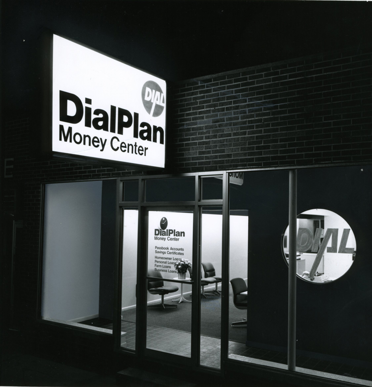 View of building entrance at night. Interior lobby seating area lights are on. Illuminated sign reads Dial Plan Money Center in black letters on white background. Image in black and white.
