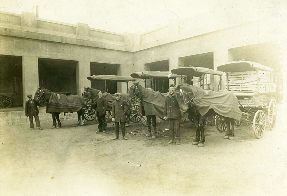 Four men are standing with four horses in a loading dock. Each horse is covered in a blanket and is hooked to a wagon.