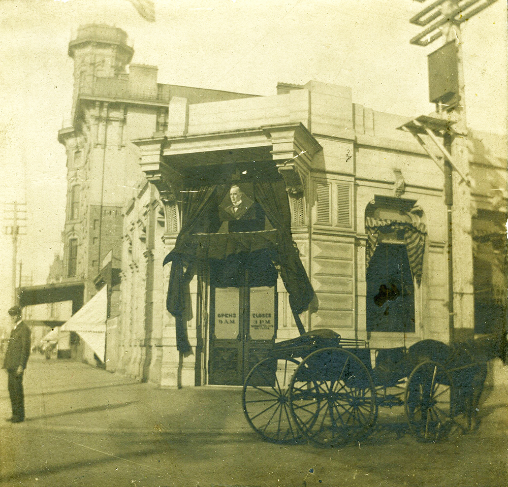 A building with black mourning crepe fabric pulled back on either side of the doors. Has a picture of President William McKinley above the doors. There is a man on the street corner and a horse drawn wagon in front of the building.