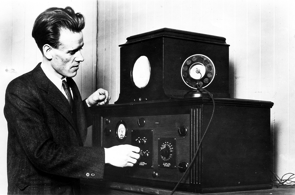 A man operates knobs on an early television, made of two wooden boxes. The larger wooden box on the bottom has dials and knobs. The smaller box has one small circular screen, smaller than the man's head.