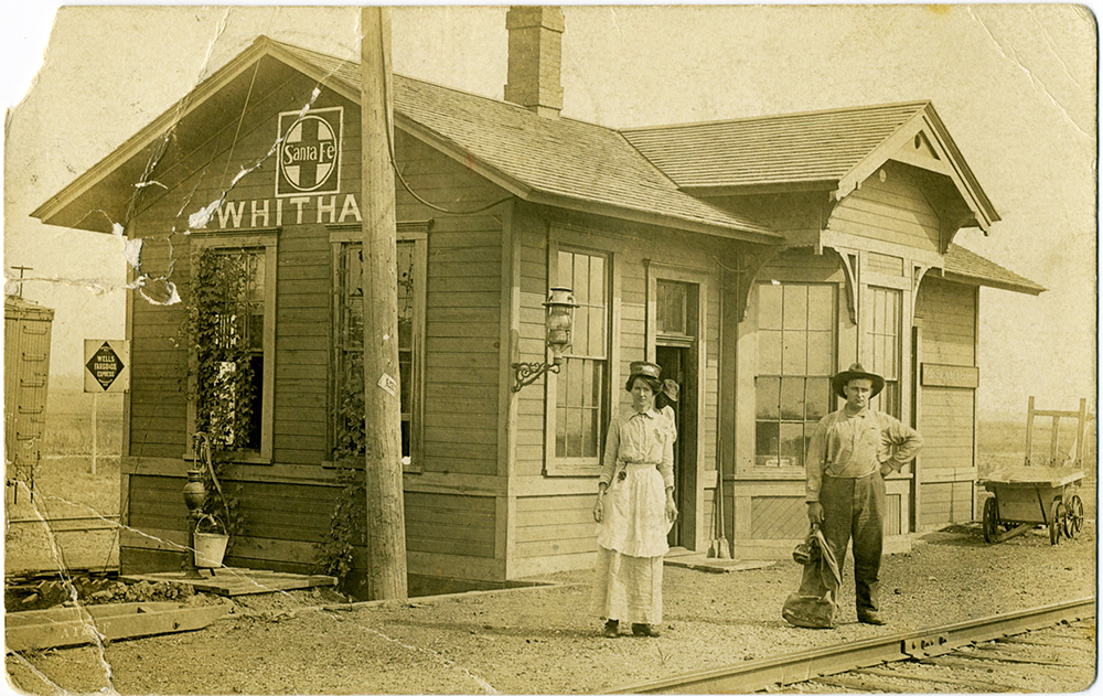 A man and a woman stand in front of a wooden building next to train tracks. Another man is seen in the doorway. The building has the name Whitham painted in white letters.