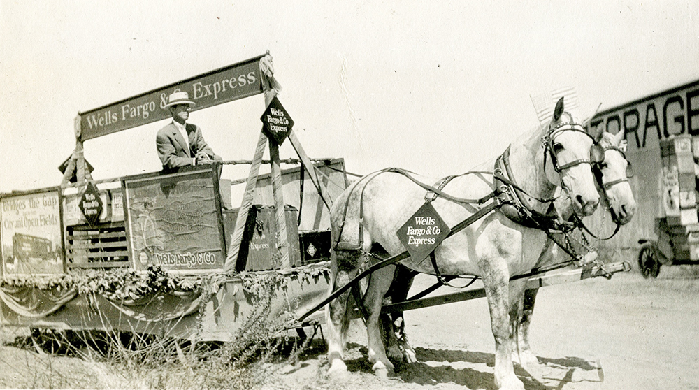 Two white horses pull a wagon as a man standing in the wagon drives them with reins. Wells Fargo and Co. Express diamond signs and banners are used for decorations.