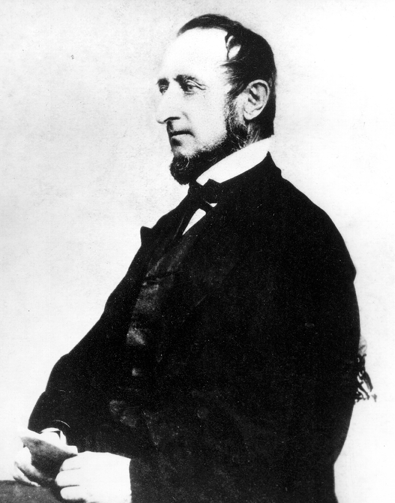 Bearded man in black suit, seated for portrait. Black and white image.