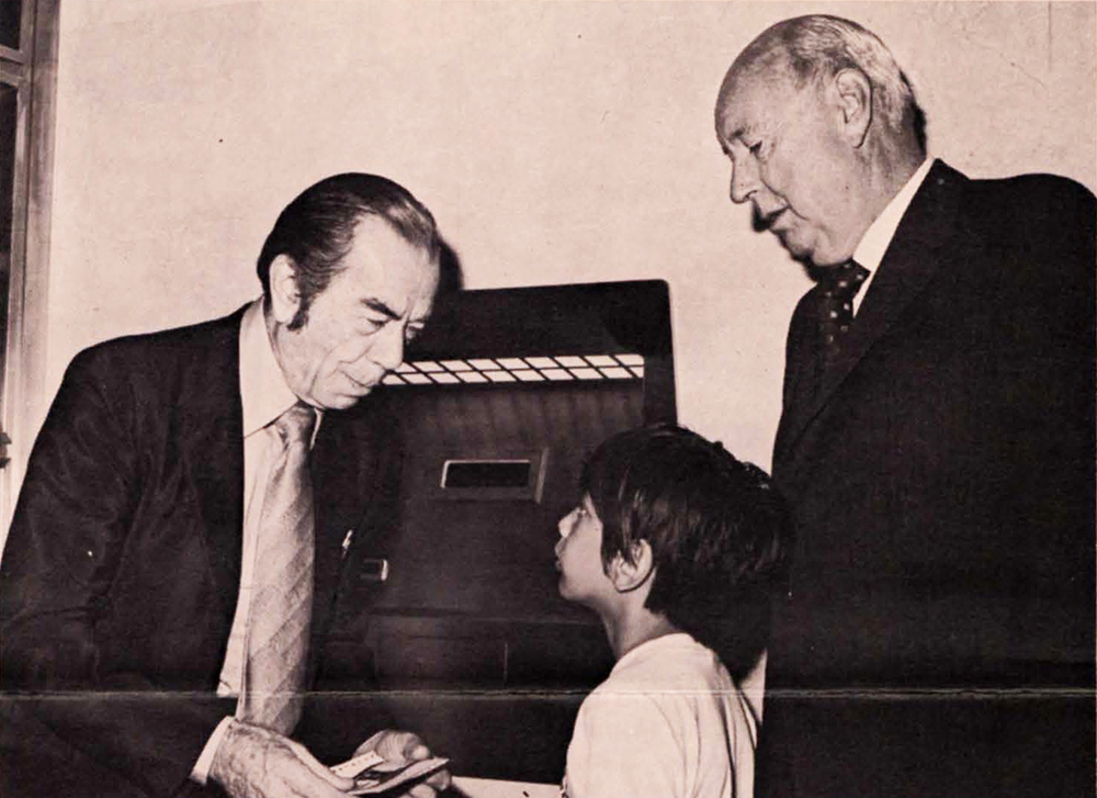 An older man in a suit and tie looks down as he speaks to a young boy who is standing in front of another older man in a suit and tie. All three are in front of an ATM. Black and white photograph.