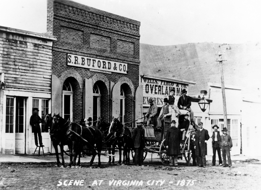 Four horses pulling a mudwagon, a type of stagecoach. Men gather nearby and on top. The stagecoach stands out front of several buildings including one with a sign: Wells Fargo and Co. Overland Mail. Handwritten label: Scene at Virginia City, 1875.