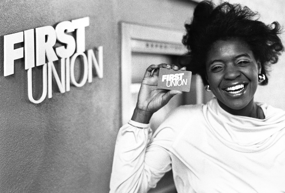 An African American woman as she smiles and holds up a First Union card. To the left is a First Union sign on the wall, and an ATM is behind her. Black and white image.