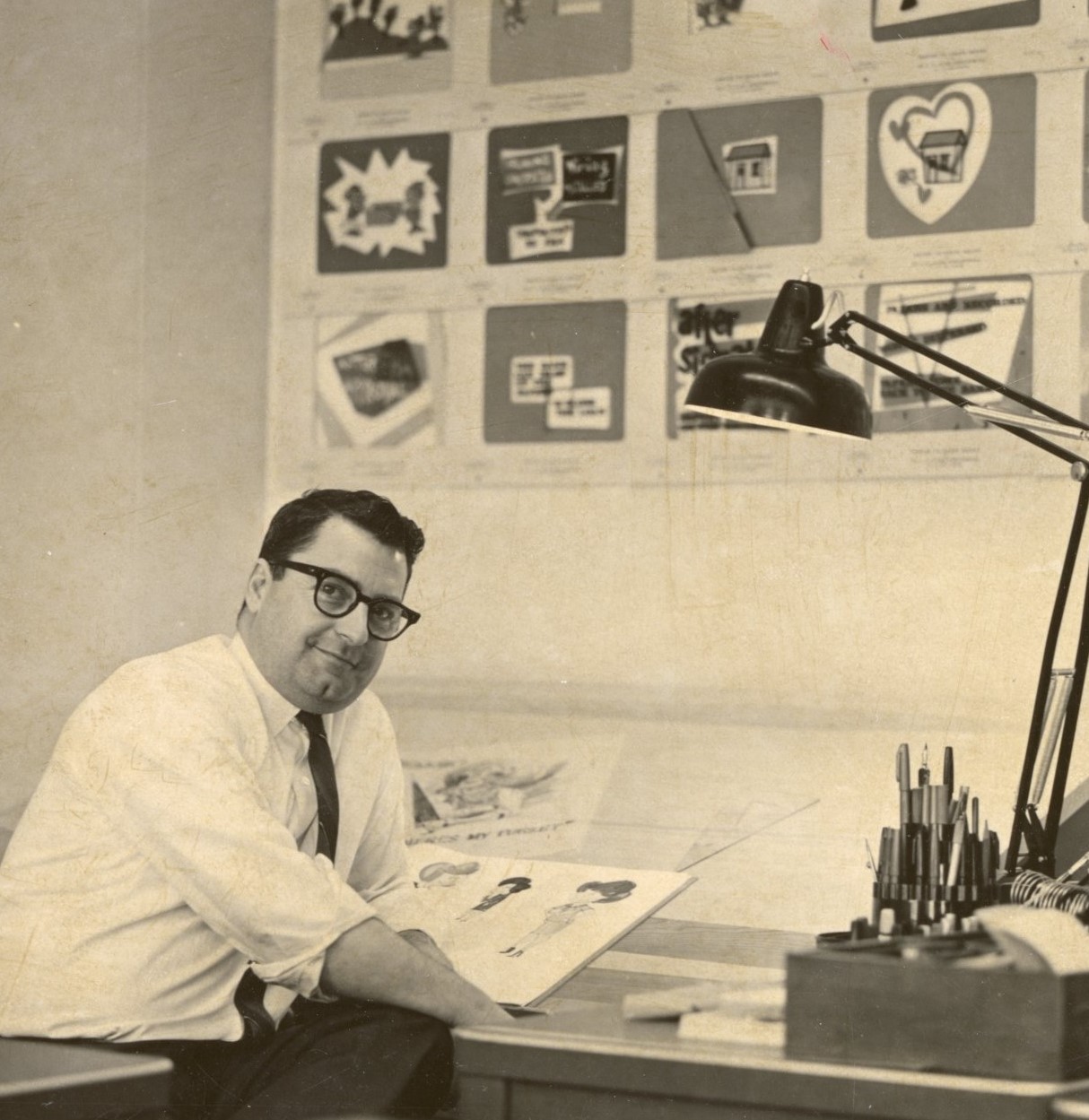 Photo of man in white shirt and tie wearing glasses sitting at artist's drafting table while looking right at camera. Cartoon art and drawings on desktop and wall behind him. Long desk lamp illuminates desk top.