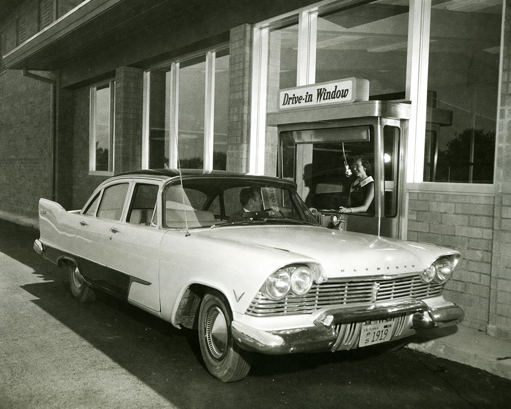 Man driving white car pulls up to brick bank building on his left. Woman teller reaches out of a window opening. A sign over head reads Drive-In Window.