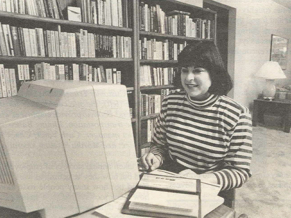 Woman sits at computer in home. On the desk is an open Wells Fargo checkbook. Behind her is a large bookshelf.