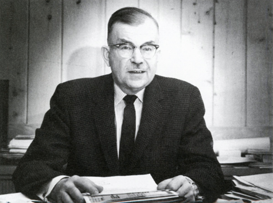 Man in black suit and tie sitting at desk holding sheaf of papers. He wears glasses and looks directly at camera. Black and white image.