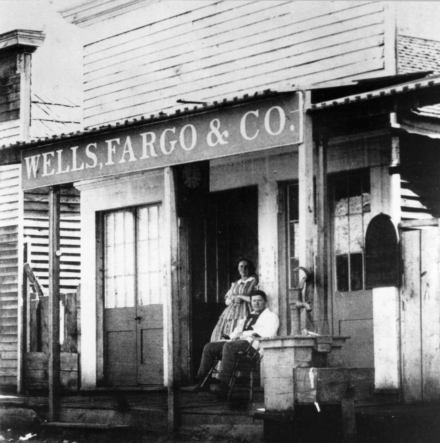 Photo of man and woman on porch. He leans back in chair wearing white shirt and dark pants. She wears long print dress and stands in doorway of small wood frame commercial building. Large sign overhead reads Wells Fargo & Co. Image is black and white.