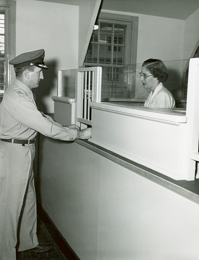 Man in military uniform stands at left at a white teller counter in a room with two windows. His hands rest on counter, under a panel of short iron bars. Opposite him, behind iron bars and low wall with glass panes is woman wearing glasses.
