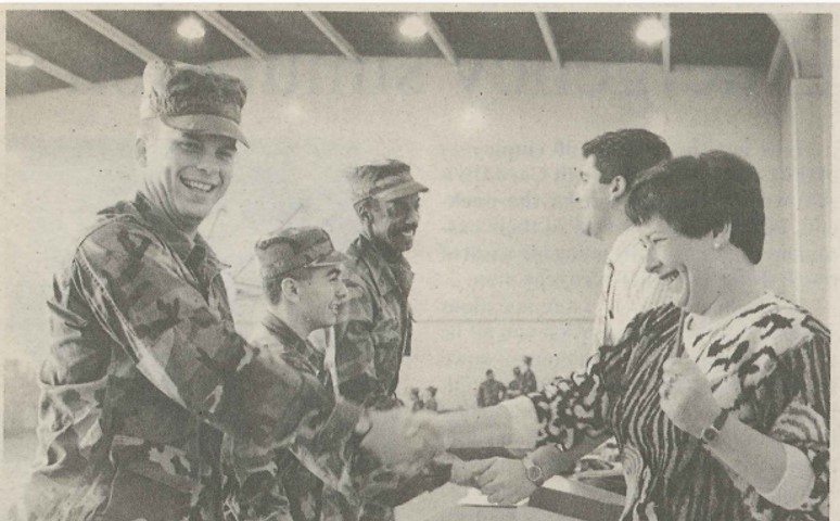 Three smiling men in military uniforms stand and shake hands over a table with two bank employees. Woman banker foreground, male banker background. Additional troops in background. Image is black and white.
