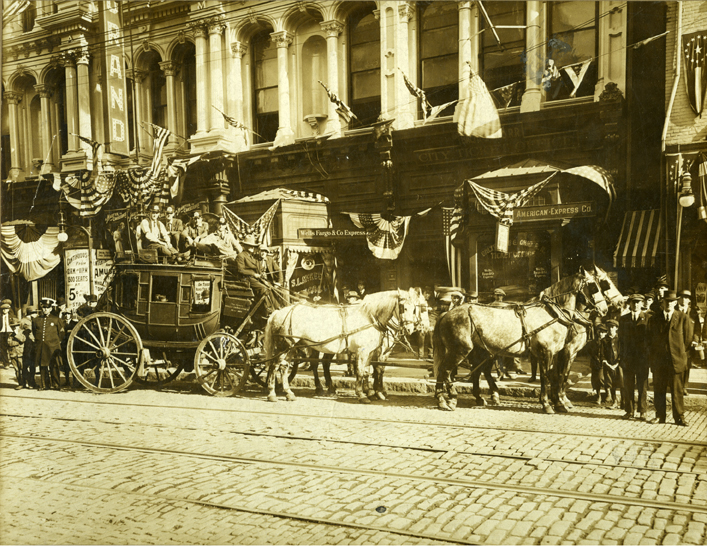 Stagecoach facing right, pulled by four horses, standing still on cobbled street. Several men stand by lead horses, at rear of coach, and sitting on top. In background, a three-story building is draped in American flag bunting. Flags fly from windows. Signs on building say GRAND, Wells Fargo & Co’s Express and American Express Company.