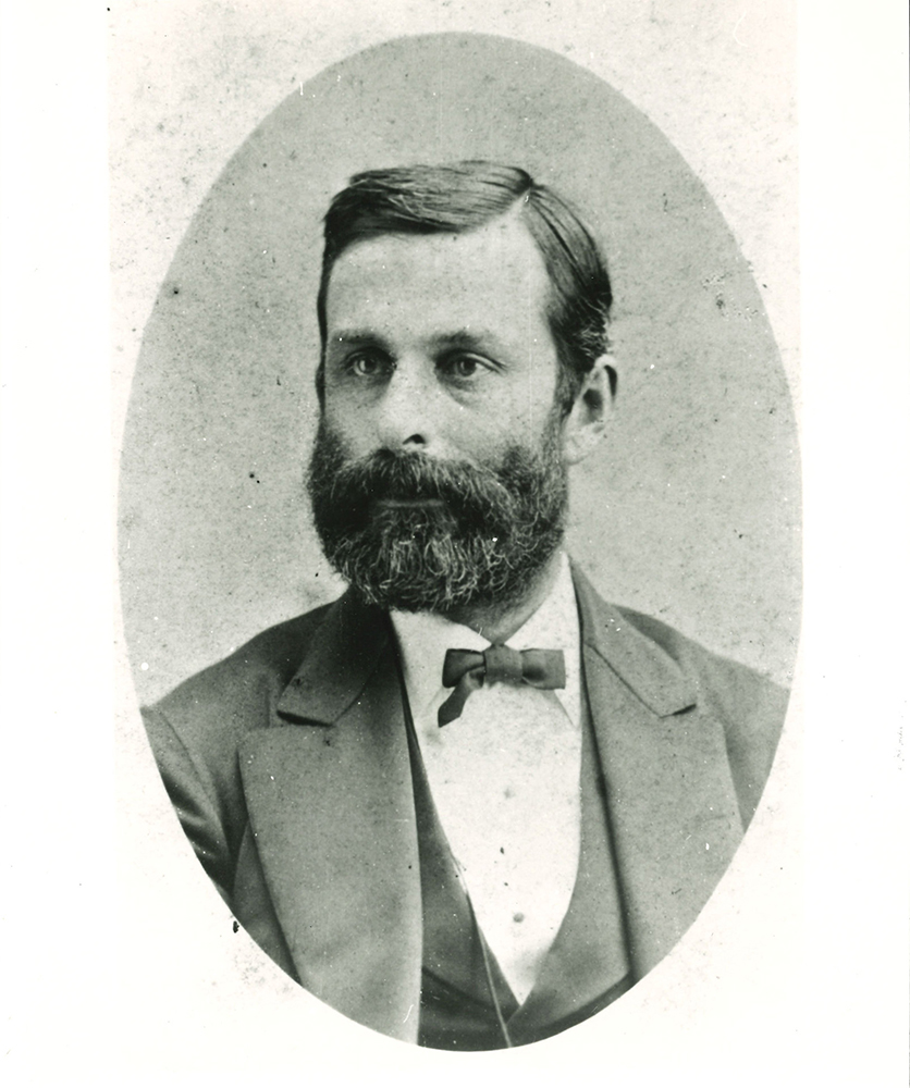 Man with beard and mustache looking right, wearing dark suit, bow tie, and vest. Image in black and white.
