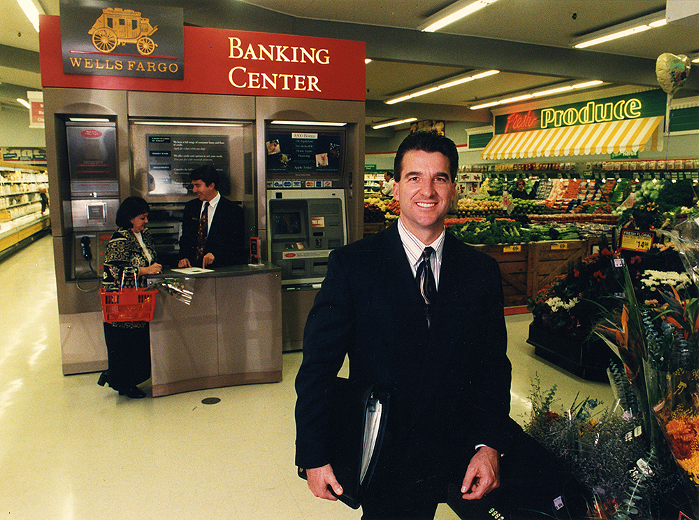 A man in suit and tie holding a binder of papers stands in front of a combination ATM unit and banking counter labeled Banking Center. A man stands behind the counter, waiting on a woman standing in front of the counter. In the background is a grocery store’s produce section.