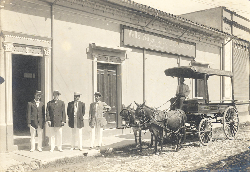 Four men in dark suit jackets, white pants and boater hats pose standing outside a white building with sign Wells Fargo & Co Express S.A. Sociedad Mexicana. An open-sided wagon facing left and drawn by two mules is at the curb to the right of the four men. A fifth man is seated in the wagon’s driver seat. The wagon is lettered Wells Fargo & Co