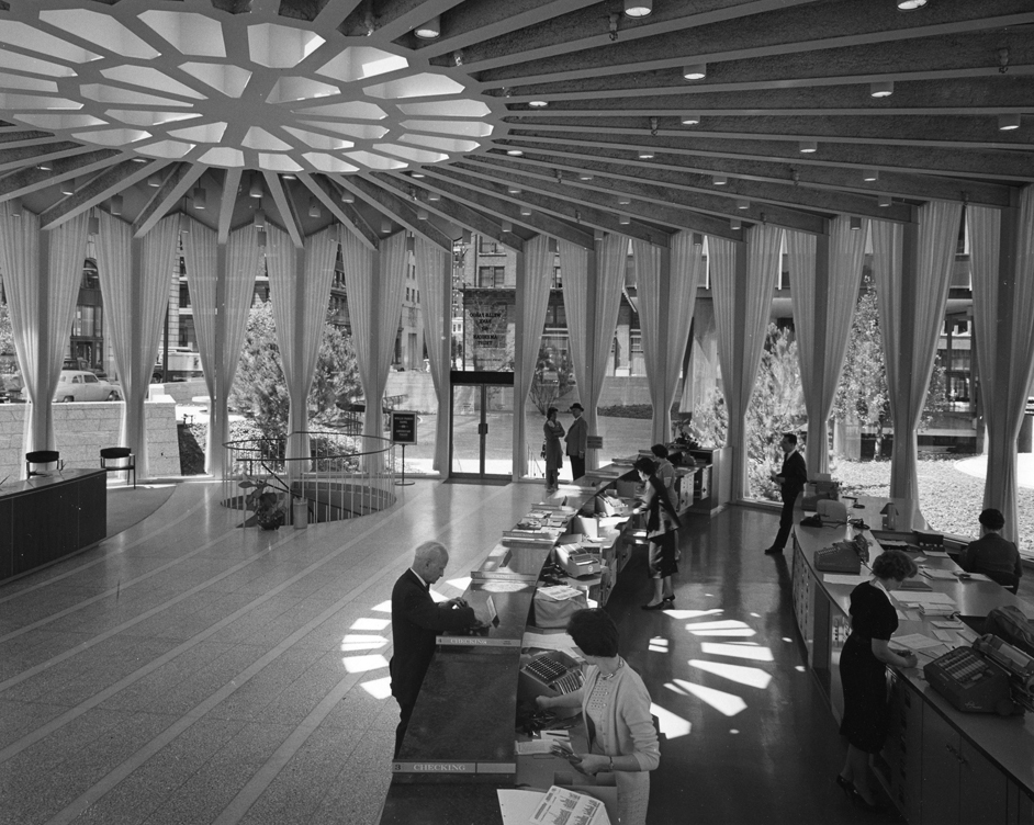 An open bank interior space, round in shape with multiple floor to ceiling windows with draperies on every side. Ceiling shows support beams radiating out in all directions from hub-like center. Natural light falls from ceiling center skylight onto floor. In the round room are people walking or standing on both sides of two low counters, also slightly rounded in crescent shape to fit the circular interior space.