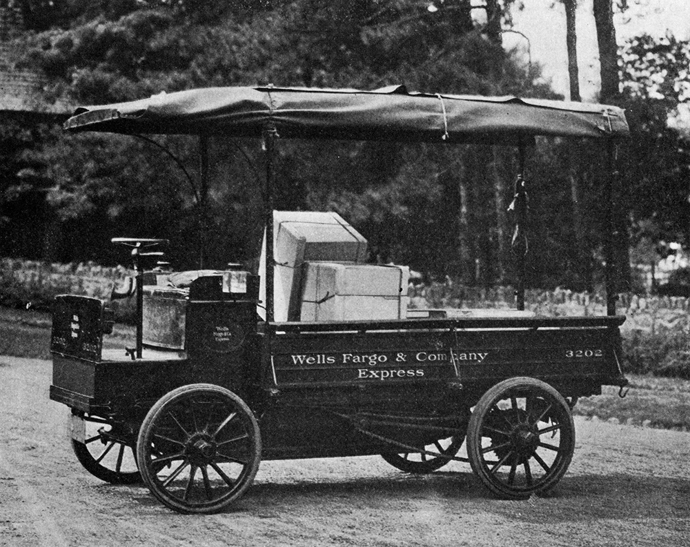 An electric truck sits on a dirt road with trees in the background. The truck is open aired with no windshield and a canvas top. The side rail reads: Wells Fargo & Company Express.