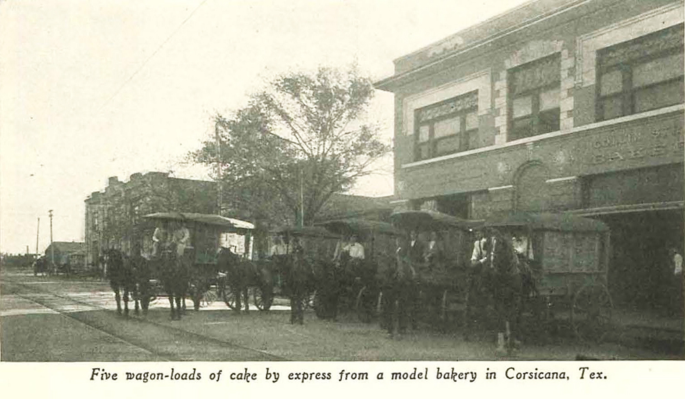 Five loaded wagons, each drawn by one or two horses, are backed to the curb in front of a two-story stone building with sign Collin Street Bakery. Several men sit on wagons or stand beside them. Photo legend says five wagon-loads of cake by express from a model bakery in Corsicana, Texas.